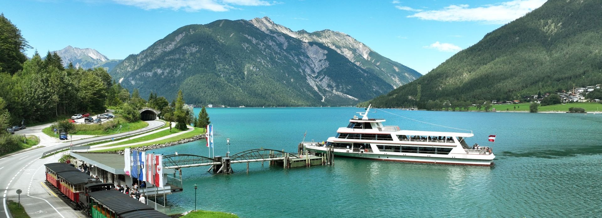 Die Achenseebahn fährt am Achensee entlang, während ein Schiff auf dem See unterwegs ist.