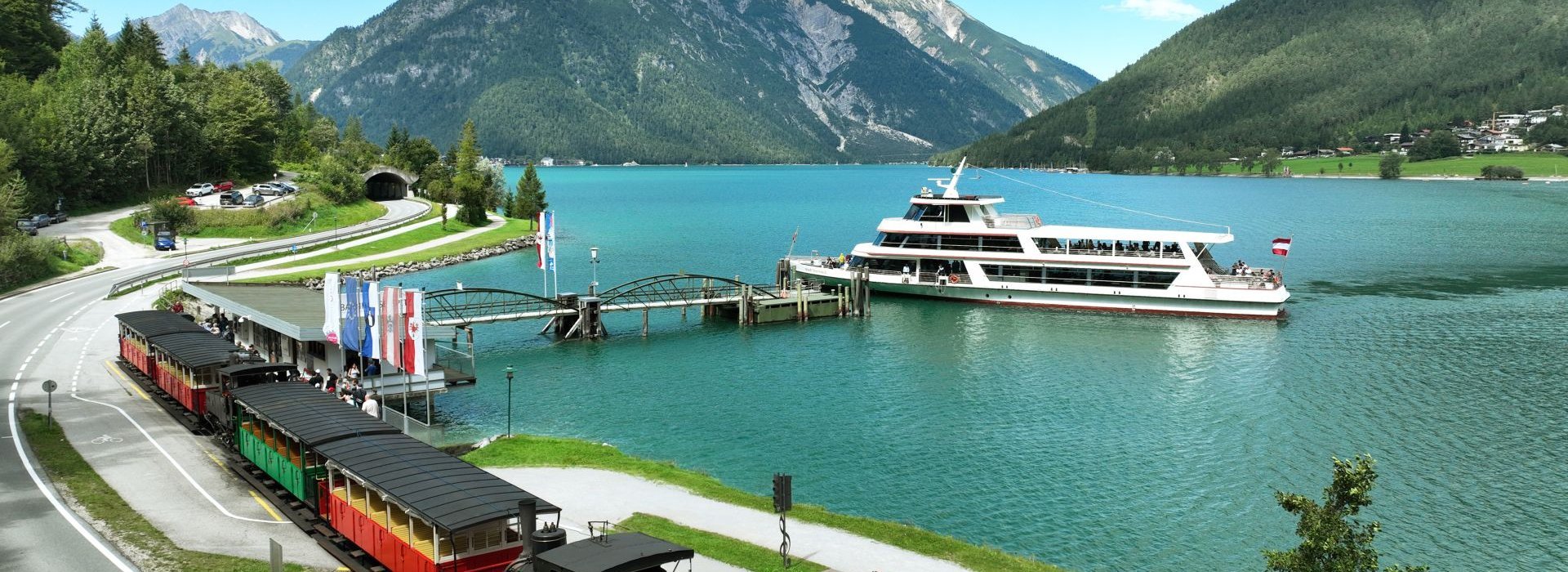 Die Achenseebahn fährt am Achensee entlang, während ein Schiff auf dem See unterwegs ist.