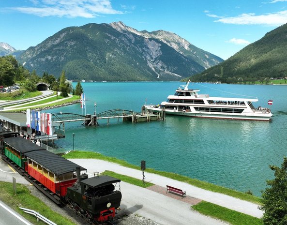 Die Achenseebahn fährt am Achensee entlang, während ein Schiff auf dem See unterwegs ist.