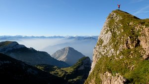 Eine Person in roter Kleidung steht auf einem Berggipfel und streckt die Hände in die Höhe.