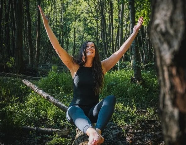 Eine lachende Frau sitzt im Wald auf einem Baumstamm.